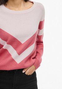 Rožinė ir šviesiai rožinė chevron rašto megztinis su apvalia iškirpte ir sutrauktomis rankovėmis, pagamintas iš minkštos medžiagos. Derinamas su juodomis džinsinėmis kelnėmis.