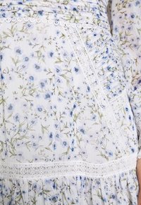 Bílá látka s malým modrým květinovým potiskem, s jemnými krajkovými lemováními a sklady na oděvu.