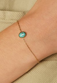 Gouden armband met een ovale turkoois steen met witte aders, geplaatst in een verguld frame, gedragen op een lichte huid.