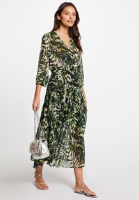 Maxikleid aus transparenter Stoff mit einem grünen und schwarzen abstrakten Muster, featuring einen V-Ausschnitt, Dreiviertelärmel und einem fließenden Rock. Silberne Handtasche.