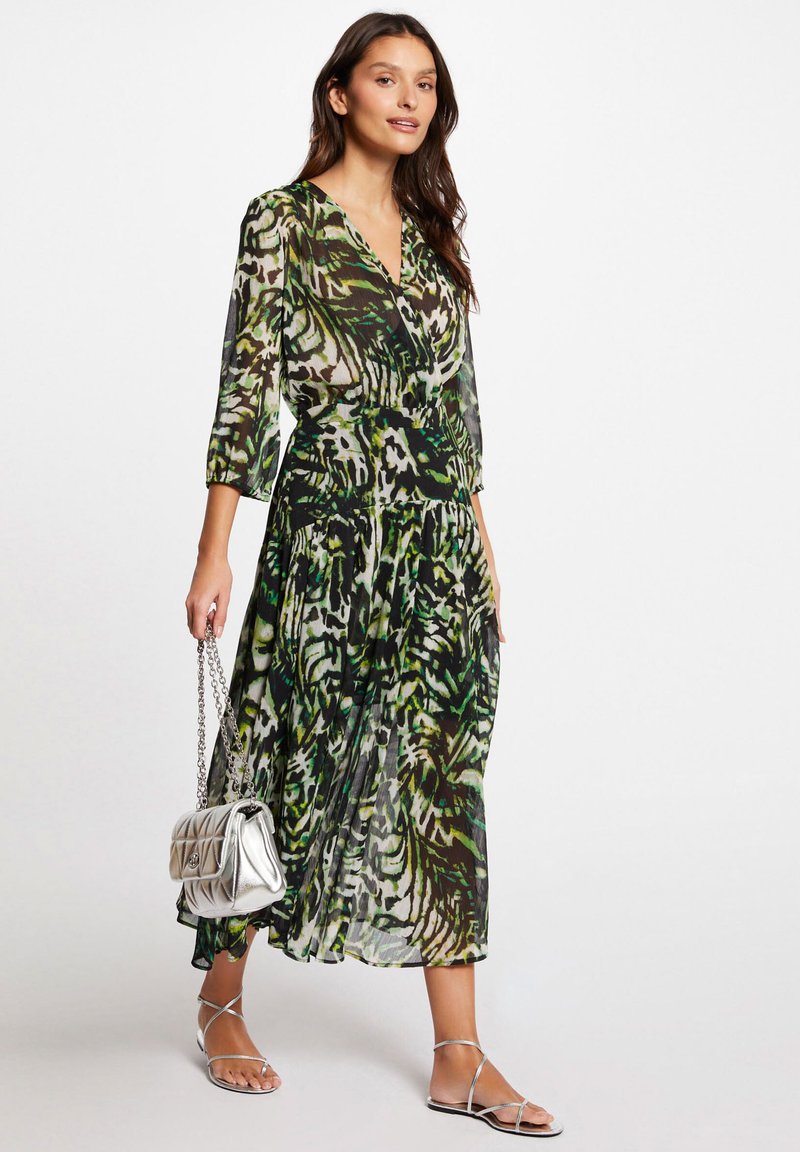 Maxikleid aus transparenter Stoff mit einem grünen und schwarzen abstrakten Muster, featuring einen V-Ausschnitt, Dreiviertelärmel und einem fließenden Rock. Silberne Handtasche.