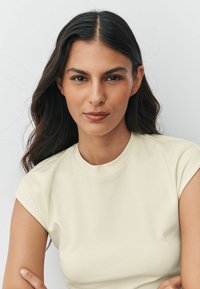 Krém színű rövid ujjú póló kerek nyakkivágással, sima anyagból készült, egy hullámos sötét hajú modell viseli.