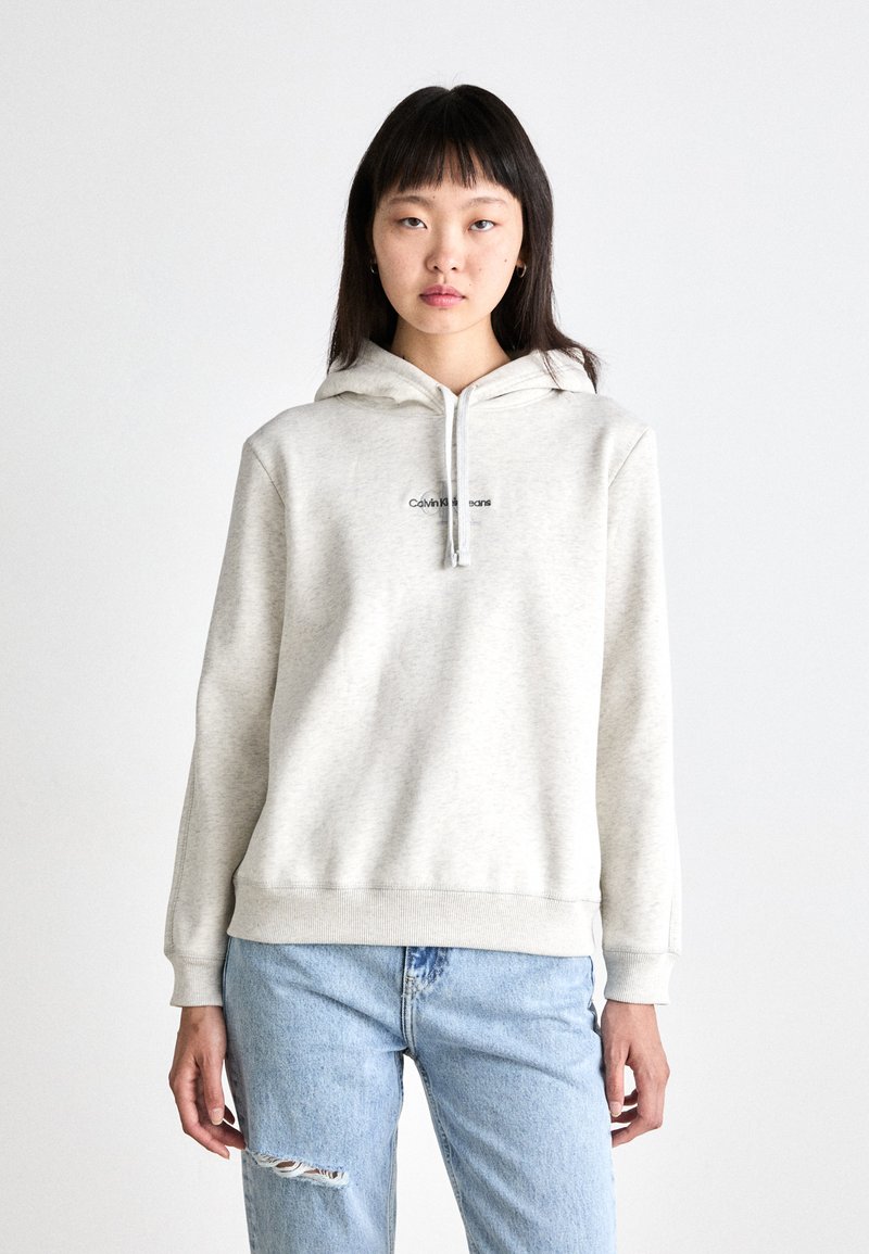Sweatshirt cinza claro com capuz, bolso frontal e cordão. Apresenta um pequeno logotipo preto no peito. Usado com jeans rasgados em azul claro.