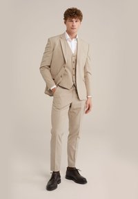 Beige Dreiteiler mit strukturiertem Schnitt, bestehend aus einem einreihigen Sakko, passender Weste und schlank geschnittenen Hosen. Dunkelbraune Schuhe.