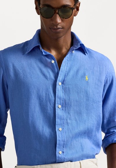 Lys blå lin-skjorte med knappelukking, hvite knapper og liten gul logo på brystet. Modellen har på seg mørke solbriller.