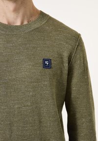 Grüner Strickpullover mit einem gerippten Rundhalskragen, der auf der Brust ein kleines blaues quadratisches Patch mit weißem Text zeigt. Weicher, strukturierter Stoff.
