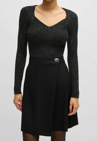 Svart stickad bodysuit med långa ärmar och V-ringning, med en texturerad mönster och silverprickar. Matchas med en svart kjol, prydd med en glänsande knapp.