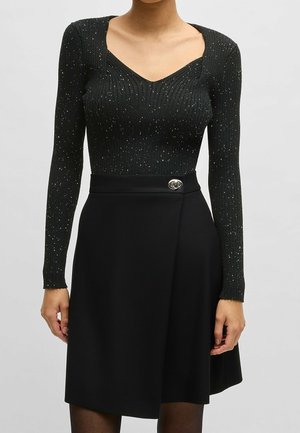 Zwarte gebreide bodysuit met lange mouwen en V-hals, voorzien van een textuurpatroon en zilveren spikkels. Gecombineerd met een zwarte rok, afgewerkt met een glanzende knop.