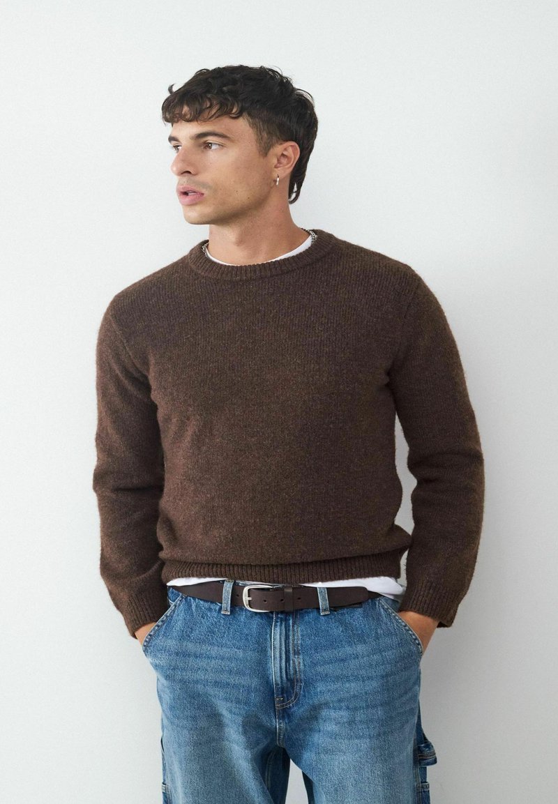 Brauner Strickpullover mit geripptem Ausschnitt und Bündchen, kombiniert mit hellblauen Jeans und einem braunen Gürtel, vor einem schlichten Hintergrund.