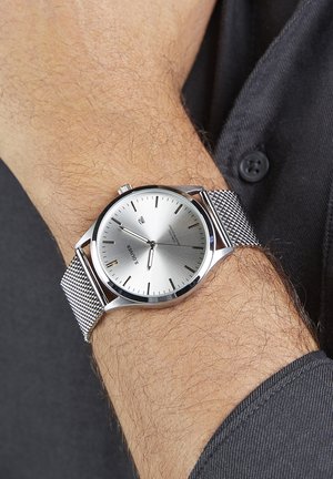 Montre à bracelet en maille argentée avec un cadran rond, cadran minimaliste argenté, aiguilles fines pour les heures et les minutes, et marquages subtils sur le bord extérieur.