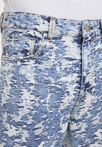 Jean en denim délavé à l'acide bleu et blanc avec des coutures visibles, un bouton en métal, et une petite étiquette de poche avant portant l'inscription "2Y STUDIOS".