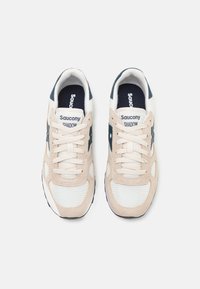 Beige és tengerészkék Saucony Shadow sportcipők, amelyek hálós és velúr anyagból készültek, kerek orrúak, fűzős kialakítással és texturált részletekkel rendelkeznek.