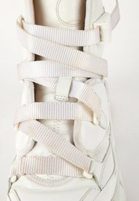 Basket blanche avec des lacets larges en satin croisés, fixés par des boucles en plastique sur un fond clair.