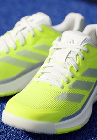 Pair of neon yellow and white running shoes with gray stripes on blue textured surface, showing mesh fabric and laces.