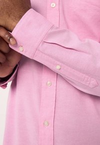 Vaaleanpunainen nappipaita, jossa on klassinen kaulus, näkyvät napit ja sileä kangaspinta, sekä taitettu hiusdetaili.