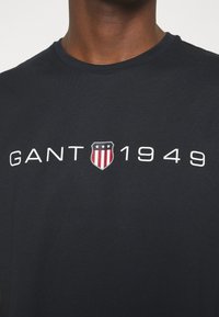 Camiseta de algodón azul marino con cuello redondo. Presenta el texto blanco "GANT 1949" y un logo de escudo con rayas rojas y blancas. Textura suave.