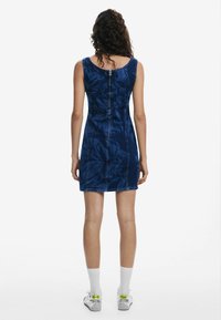 Rochie din denim de culoare albastru închis, cu un model texturat de frunze, design fără mâneci și închidere cu fermoar la spate. Purtați cu șosete albe și adidași.