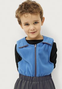 Blå fleece vest med lynlås foran, med skulderstropper. Bærer den over en sort langærmet skjorte, parret med grå elastiske bukser med talje.