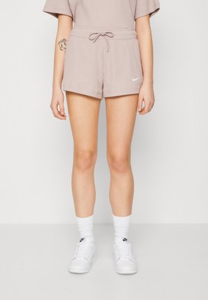 Personne portant un short côtelé beige avec un cordon de serrage, des chaussettes blanches Nike et des baskets blanches Nike, se tenant devant un fond uni.