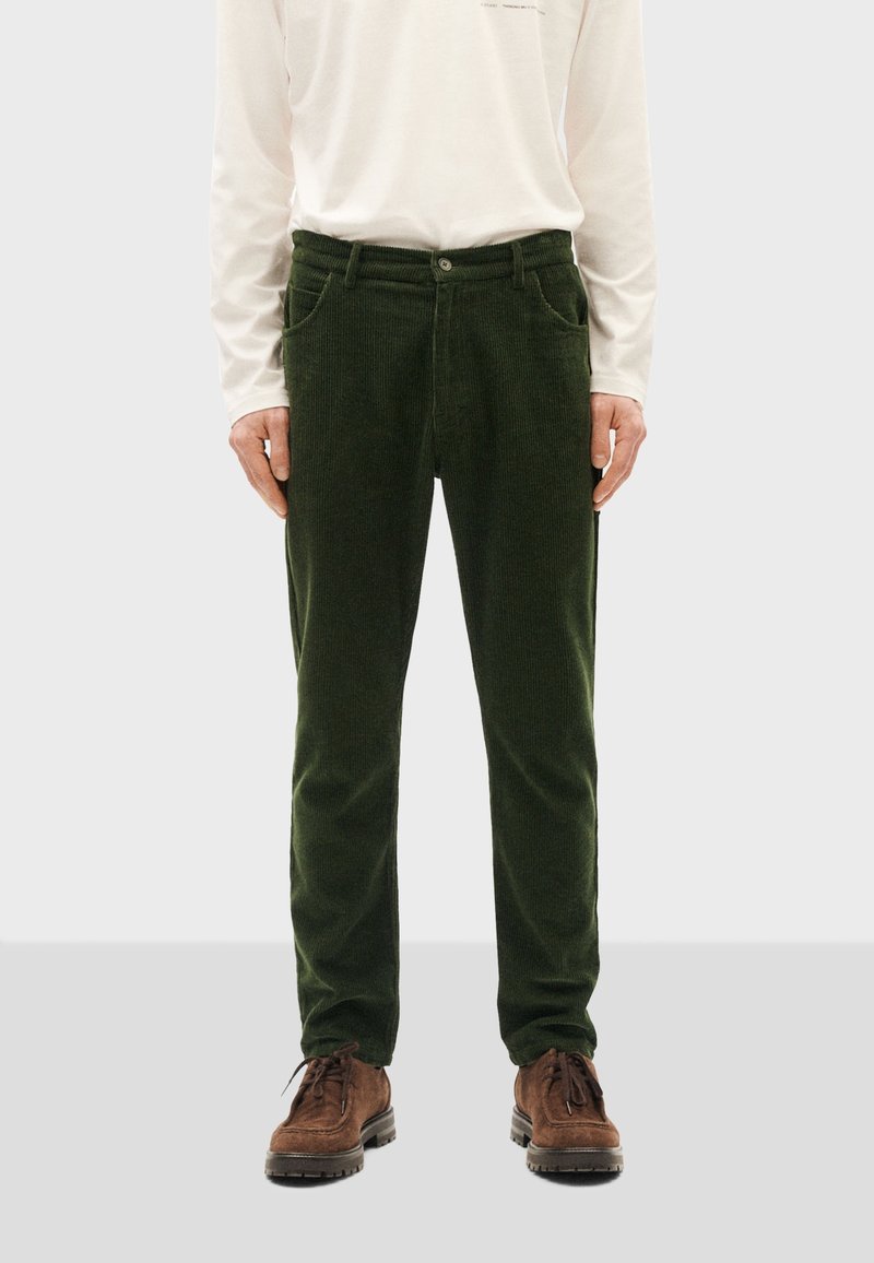 Homme portant un pantalon en velours côtelé vert foncé, une chemise blanche à manches longues rentrée, et des chaussures marron à lacets, debout sur un fond uni.