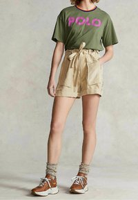 Olive green t-shirt with "POLO" in purple lettering, paired with beige high-waisted shorts featuring pockets and a tied waist, worn with brown sneakers.