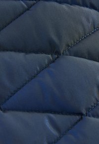 Donkerblauwe doorgestikte stof met een gladde textuur. Voorzien van diamantpatronen en contrasterende stiksels. Reflecterende glans zichtbaar op het oppervlak.