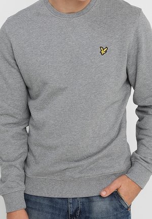 Mann trägt ein schlichtes graues Sweatshirt mit Rundhalsausschnitt und kleinem gelben Vogel-Logo auf der linken Brust, kombiniert mit blauen Jeans.