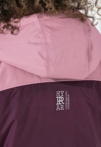 Lockiges Haar und Kapuze einer zweifarbigen lila-pinken Jacke mit dem Logo "HYR AR Outstanding Outerwear" auf der Rückseite.