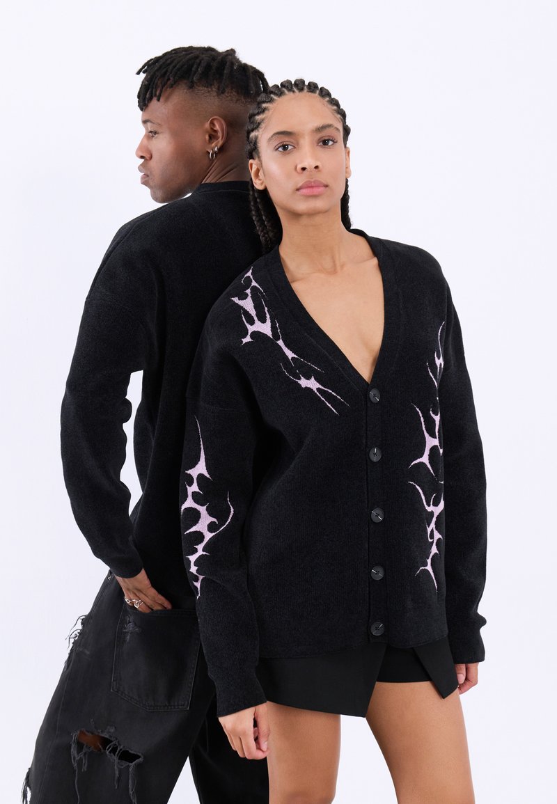 Μαύρο καμπαρντίνα με V-λαιμόκοψη, υφασμάτινη υφή, με ροζ φλόγες στα μανίκια και μαύρα κουμπιά. Συνδυάζεται με μαύρα παντελόνια και σορτς.