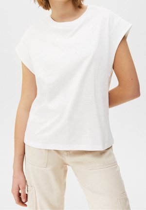 Persoon draagt een effen wit mouwloos topje en lichtbeige broek met grote voorzakken tegen een effen achtergrond.