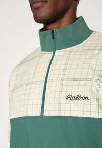 Πουλόβερ με φερμουάρ στο ένα τέταρτο, σε εκρού και πράσινο χρώμα, με μοτίβο καρό, ριμπ γιακά κ αι κεντημένο λογότυπο στο στήθος με νήμα σε ταιριαστό πράσινο.