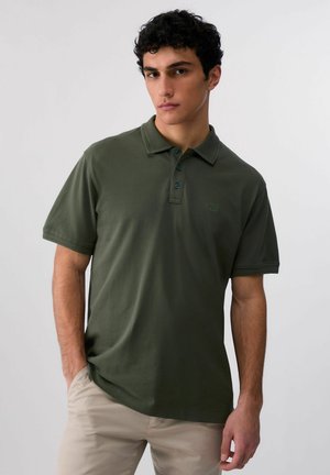 Giovane uomo con capelli ricci neri, indossa una polo verde oliva a maniche corte e pantaloni beige, in piedi con una mano in tasca.