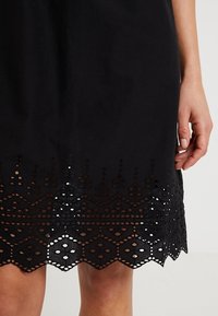 Černé šaty s vlnkovaným lemem a jemnými výřezovými vzory. Jsou vyrobeny z lehkého materiálu, mají hladkou texturu a přiléhavý střih.