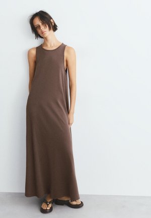 Jonge vrouw met kort donker haar, die een mouwloze bruine maxi-jurk en zwarte sandalen draagt, staand tegen een simpele witte muur.