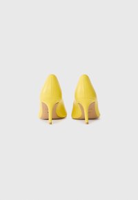 Paire de chaussures à talons jaunes montrée de dos sur un fond gris clair uni.