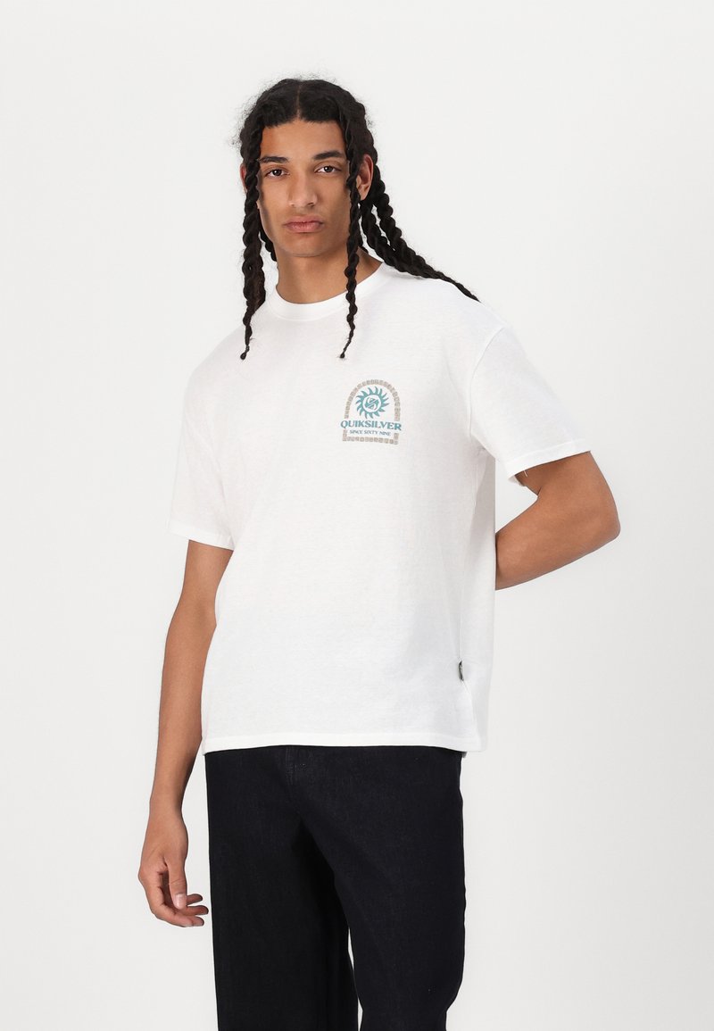 T-shirt bianco in cotone con logo grafico blu stampato sul petto. Collo a giro e vestibilità comoda, abbinato a pantaloni scuri.