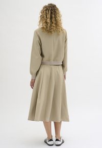 Rochie bej, cu mânecă lungă, având o talie ajustată și fustă pliată, confecționată dintr-un material neted. Purtați cu adidași negri și albi.