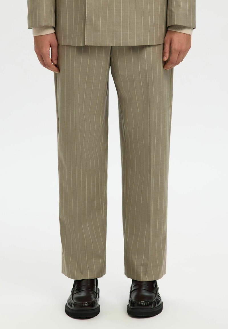 Homme portant un pantalon de costume beige à fines rayures, avec un blazer assorti et des mocassins en cuir noir, debout sur un fond blanc.