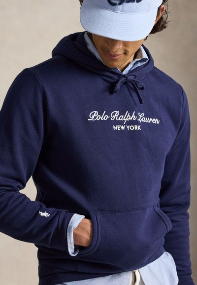 Άτομο που φοράει σκούρα μπλε φούτερ Polo Ralph Lauren με λογότυπο κειμένου και ανοιχτό μπλε καπέλο, με το χέρι στην μπροστινή τσέπη του φούτερ.