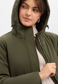 Veste vert olive avec capuche, dotée d'une fermeture éclair à l'avant, de coutures discrètes et d'une texture douce. Portée sur un haut de couleur claire.