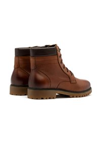 Brązowe skórzane botki za kostkę o gładkiej fakturze, z przodu sznurowane i z brązową gumową podeszwą. Posiadają kontrastujące czarne wykończenia przy kostce oraz metalowe akcenty.