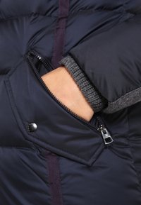 Marineblauwe donzen jas met een gestructureerde grijze gebreide manchetten, een ritssluiting met drukknop en contrasterende donkerpaarse accenten.