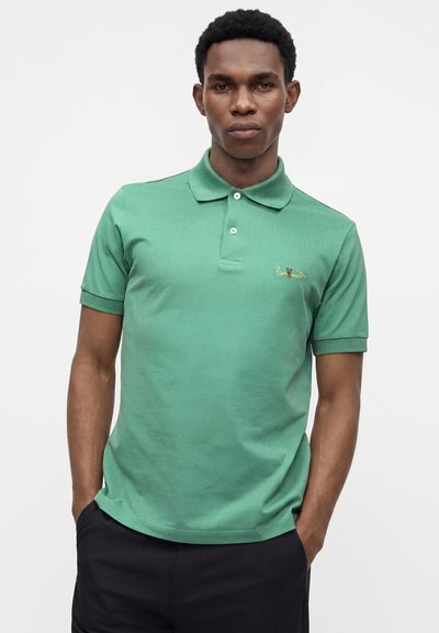 Homme portant un polo vert à manches courtes avec le logo Paul Smith, debout les mains dans les poches devant un fond uni.