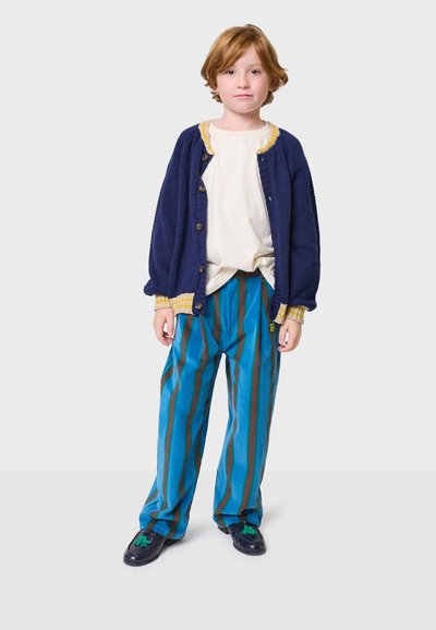 Mornarska pletena cardigan s žutim rubom, bijela košulja, plave i smeđe prugaste hlače te crne cipele s zelenim akcentima. Dijete stoji uspravno.