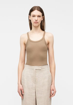Femme portant un débardeur beige ajusté et un pantalon en lin beige clair taille haute, debout devant un fond clair uni.