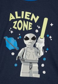 Navyblaues T-Shirt mit einem Grafikdesign eines grauen Aliens, das ein Lichtschwert hält, beschriftet mit "ALIEN ZONE", sowie Akzentsternen und -formen in verschiedenen Farben.