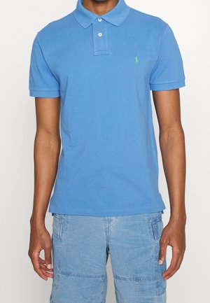 Man draagt een blauw poloshirt met korte mouwen, kraag en knopen, en lichtblauwe cargoshorts met meerdere zakken.