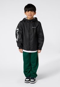Ragazzino in piedi e sorridente, con una giacca Champion nera, pantaloni sportivi verdi e sneakers bianco e nere su uno sfondo bianco.