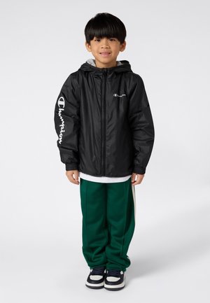Ragazzino in piedi e sorridente, con una giacca Champion nera, pantaloni sportivi verdi e sneakers bianco e nere su uno sfondo bianco.
