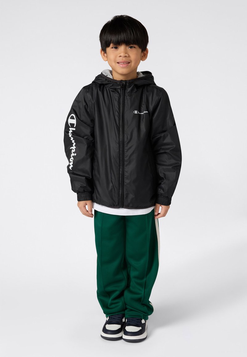 Ragazzino in piedi e sorridente, con una giacca Champion nera, pantaloni sportivi verdi e sneakers bianco e nere su uno sfondo bianco.